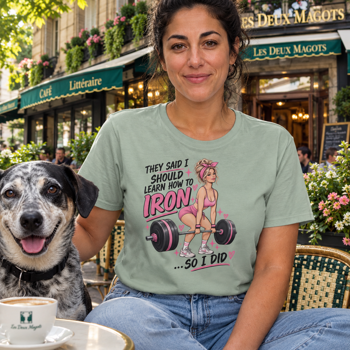 A woman wearing a sage green graphic t-shirt sits at an outdoor cafe table next to a black and white dog. The t-shirt features a graphic of a woman lifting a barbell with the text 'THEY SAID I SHOULD LEARN HOW TO IRON .