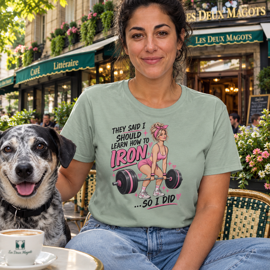 A woman wearing a sage green graphic t-shirt sits at an outdoor cafe table next to a black and white dog. The t-shirt features a graphic of a woman lifting a barbell with the text 'THEY SAID I SHOULD LEARN HOW TO IRON .